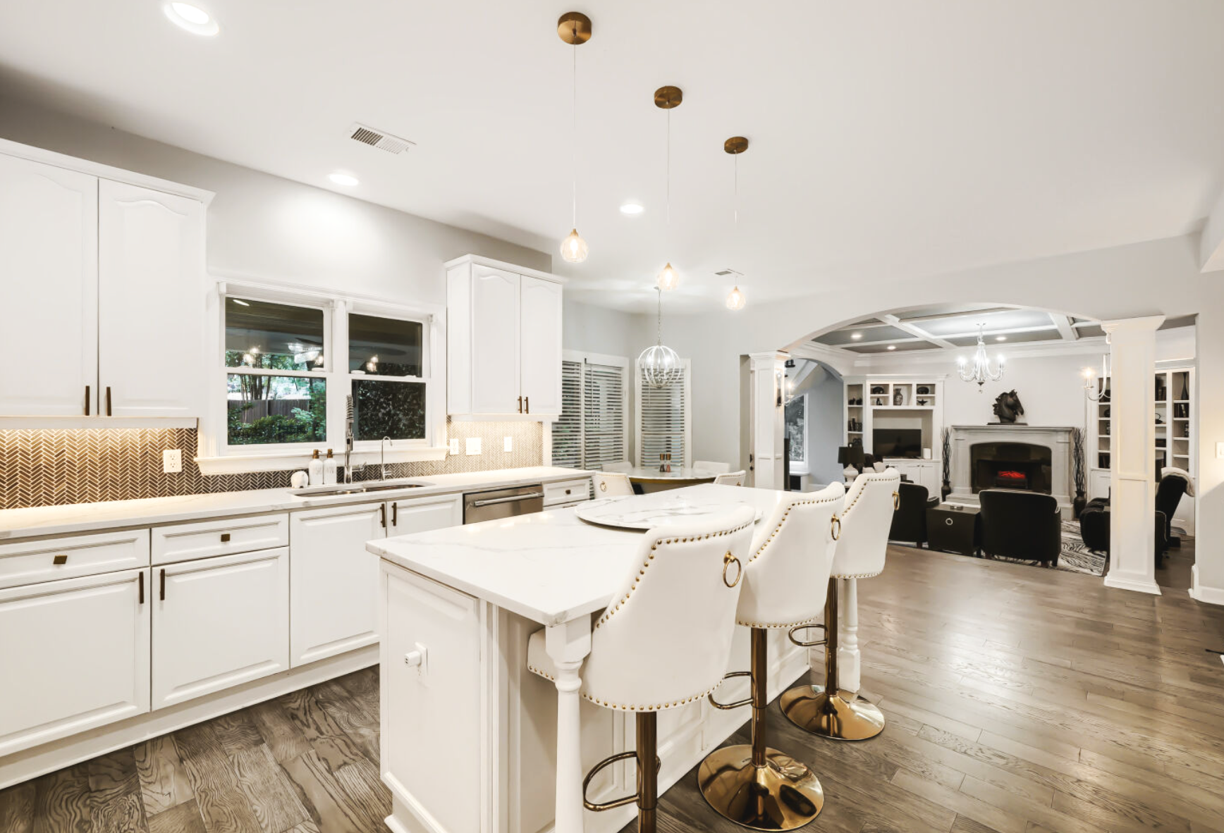 Modern white kitchen with island, bar stools, and view into living room with fireplace and seating area.
