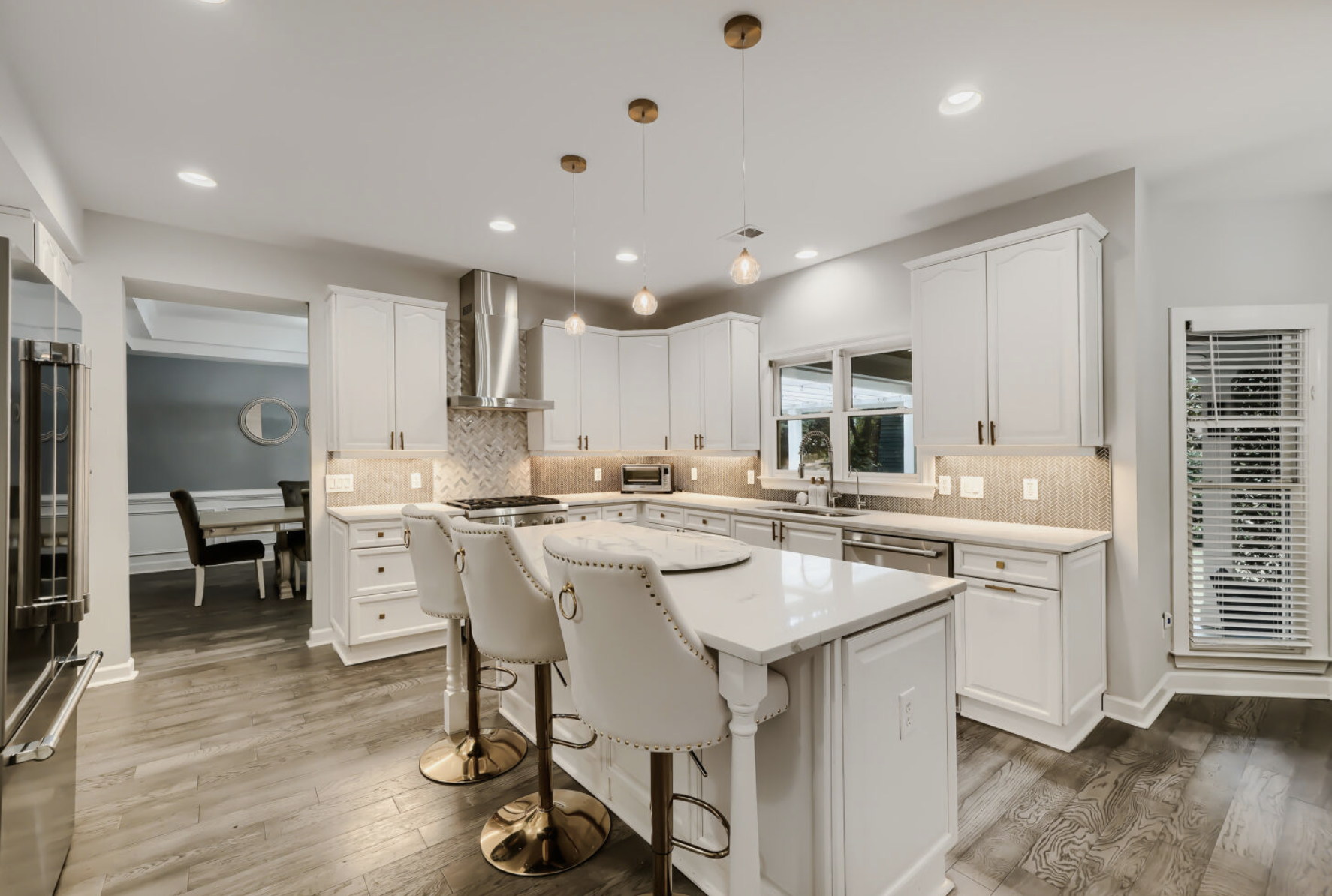Modern white kitchen with island, pendant lights, stainless steel appliances, and a small dining area visible through a doorway.