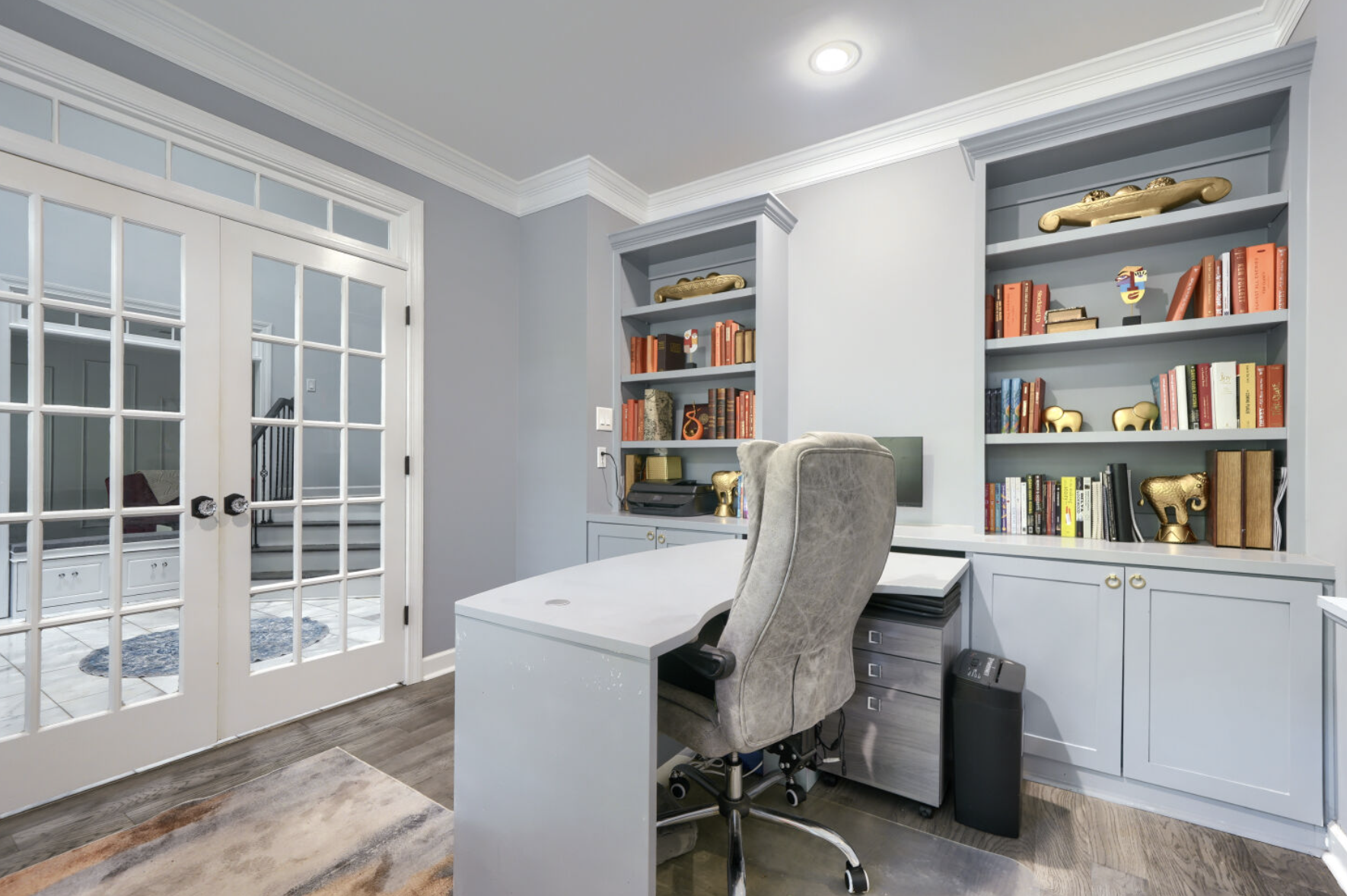 A home office with light gray walls, white cabinetry, and built-in bookshelves filled with books and decorative items, including gold animal figurines and art pieces. There is a large desk with a gray high-back office chair, a computer monitor, and a printer. French doors with glass panes lead to an adjacent room, and a rug with abstract patterns is on the wooden floor.