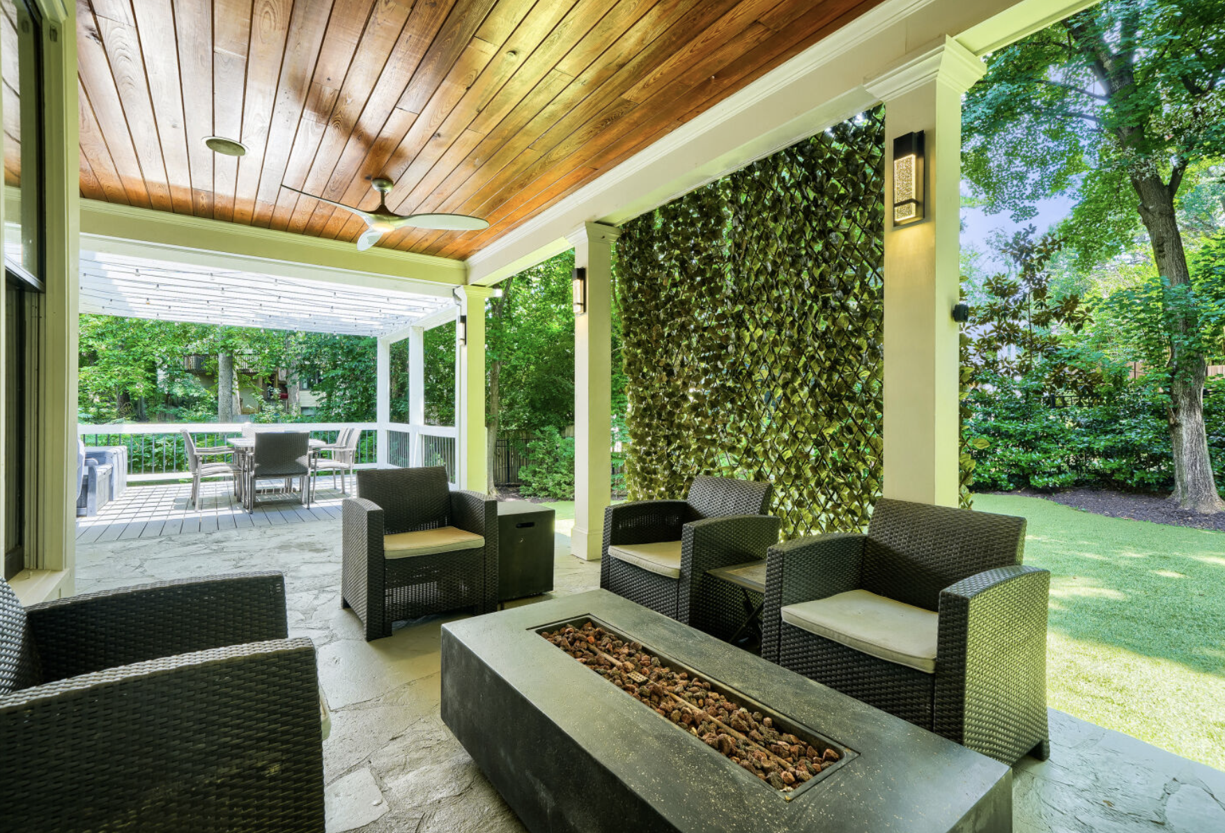 Covered patio with wicker chairs, a fire pit, and a deck with a table and chairs in a lush green backyard.
