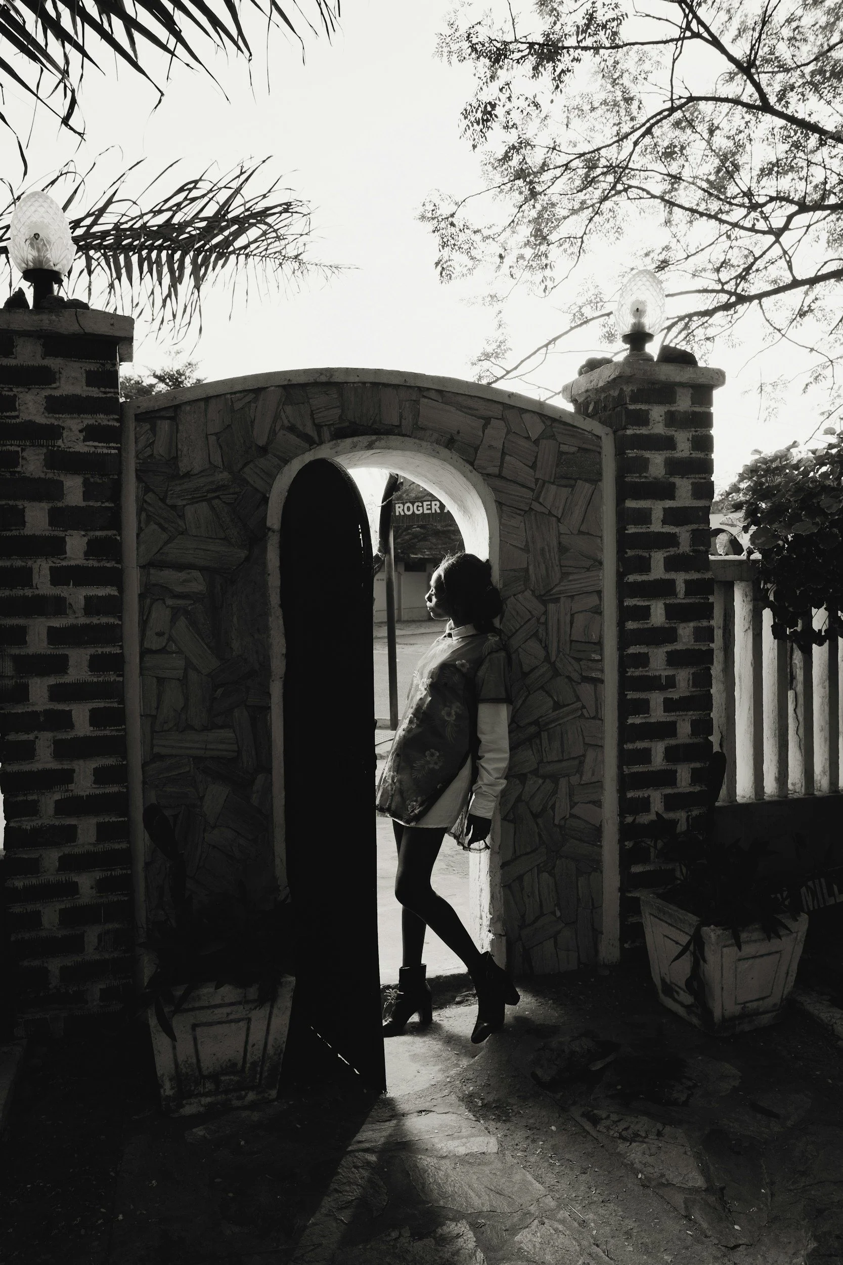 A black and white photo of a woman with curly hair standing under an archway made of stone and wood. She is wearing high-heeled boots, shorts, and a jacket, with her hands in her pockets, looking slightly to the right. The archway is part of a brick and stone structure, with two lamps on top of the pillars. The background shows trees and a street sign reading 'ROGER'.