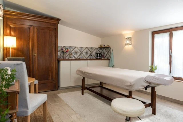 A relaxing room with a massage table, wooden furniture, a small kitchenette, and a window with curtains.