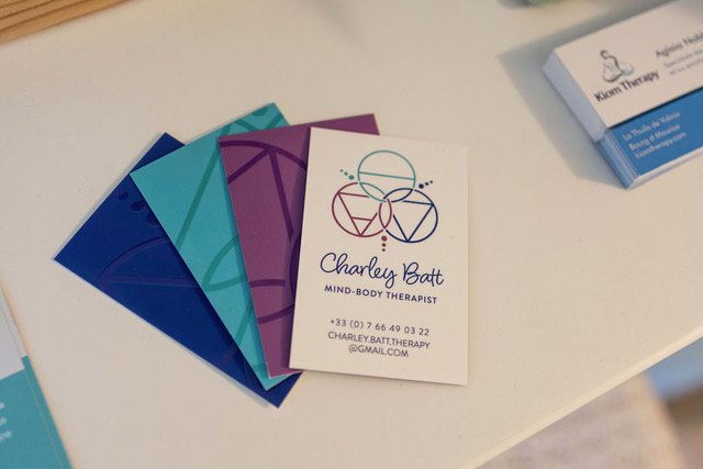 Business card for Charley Batt, a mind-body therapist, with colorful abstract logo and contact information, placed on a white surface alongside colorful folders.