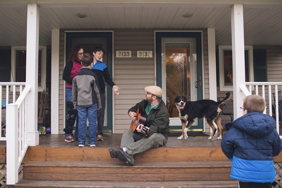 Group of people on a porch, with one playing guitar, others singing or listening, and a dog nearby.