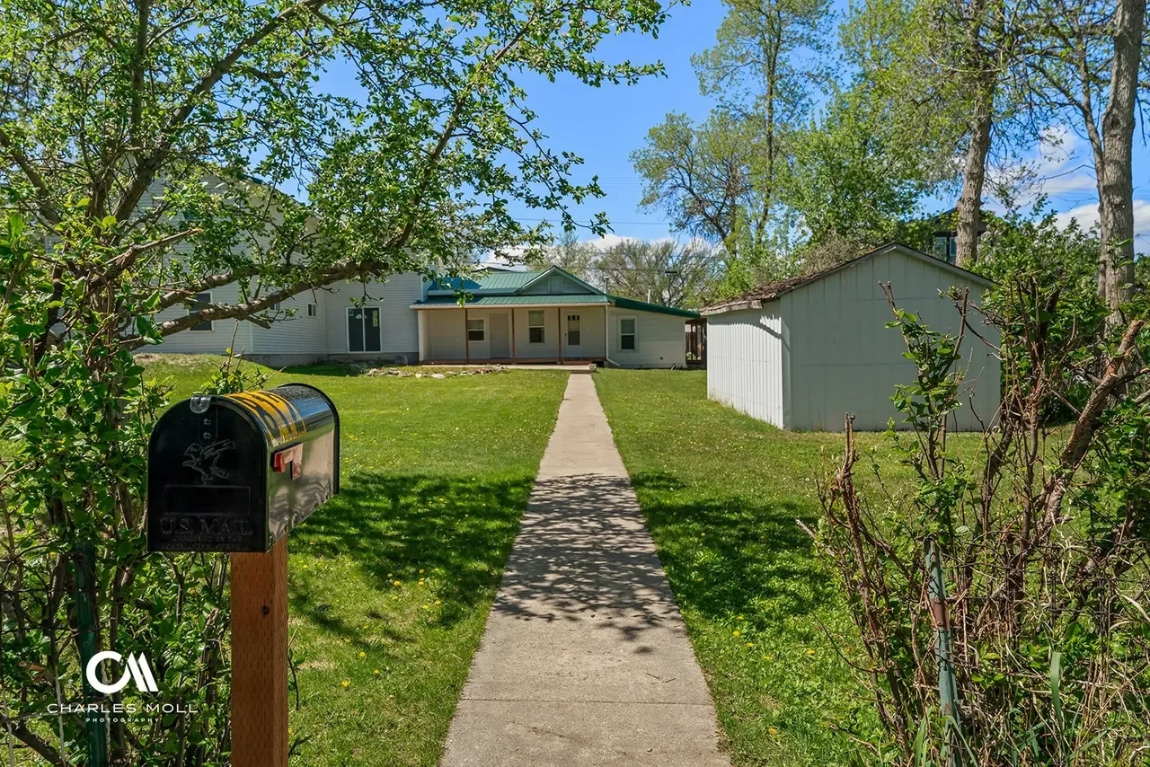 A sidewalk leading to a white house with a porch, surrounded by green trees and lawn, with a black mailbox on a wooden post in the foreground.