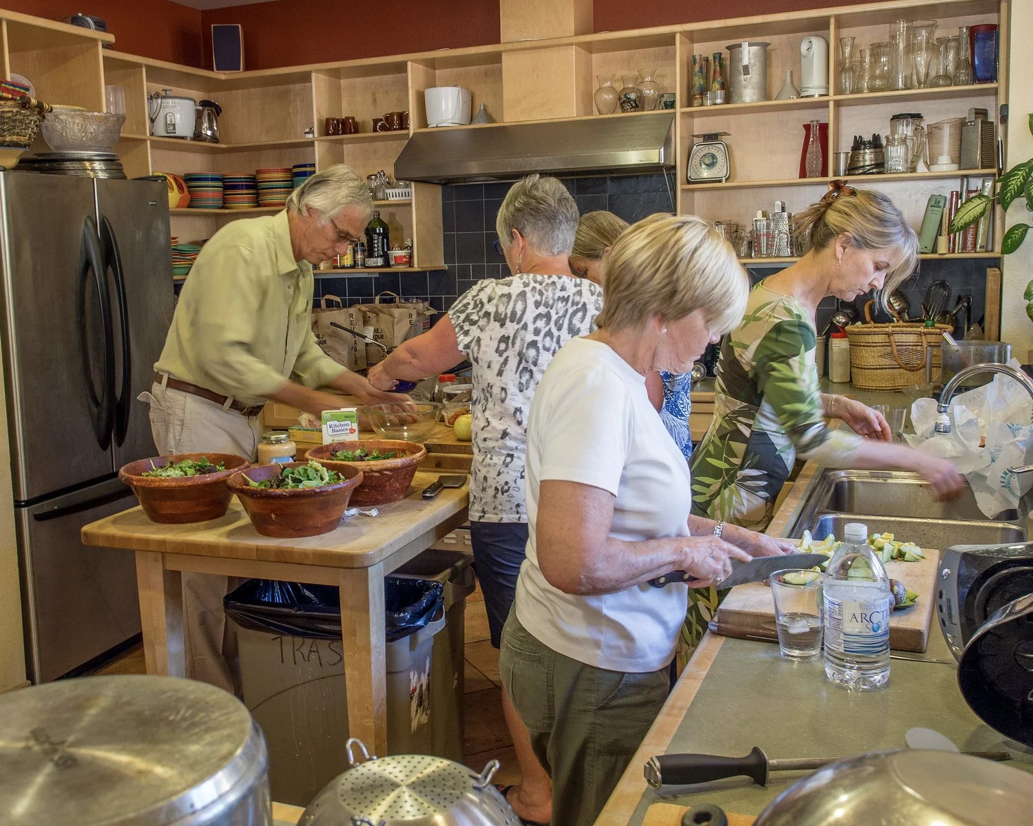 A group of five women and one man preparing food in a kitchen, with bowls of salad, sliced vegetables, and various kitchen tools on the counters.