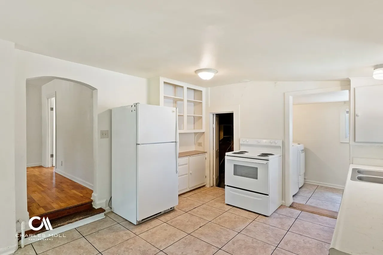 Empty kitchen with white appliances, including a refrigerator and stove, beige tile flooring, and white cabinets. A doorway leads to a laundry area with a washing machine. The walls are white, and there is a simple ceiling light.