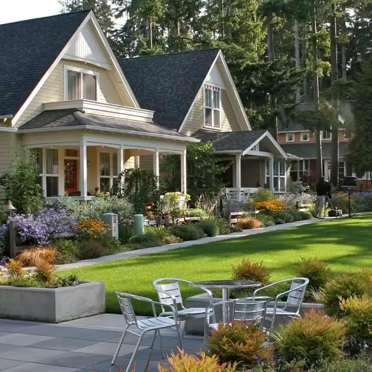A charming residential neighborhood with houses featuring gabled roofs, surrounded by lush greenery and well-maintained gardens. Outdoor patio furniture is visible in the foreground.