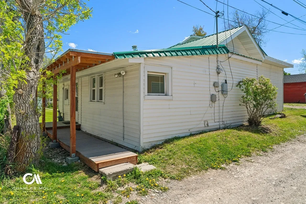 Backside of a small white house with a green metal roof, a small porch with a wooden ramp, and a tree on the left side. The yard has grass and scattered yellow dandelions, with a gravel path next to the house. Clear blue sky with a few clouds.