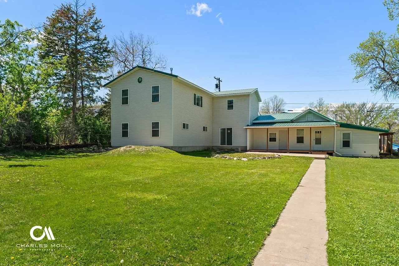 A two-story white house with multiple windows, a metal roof, a porch on the right side, surrounded by a green lawn and trees under a clear blue sky.