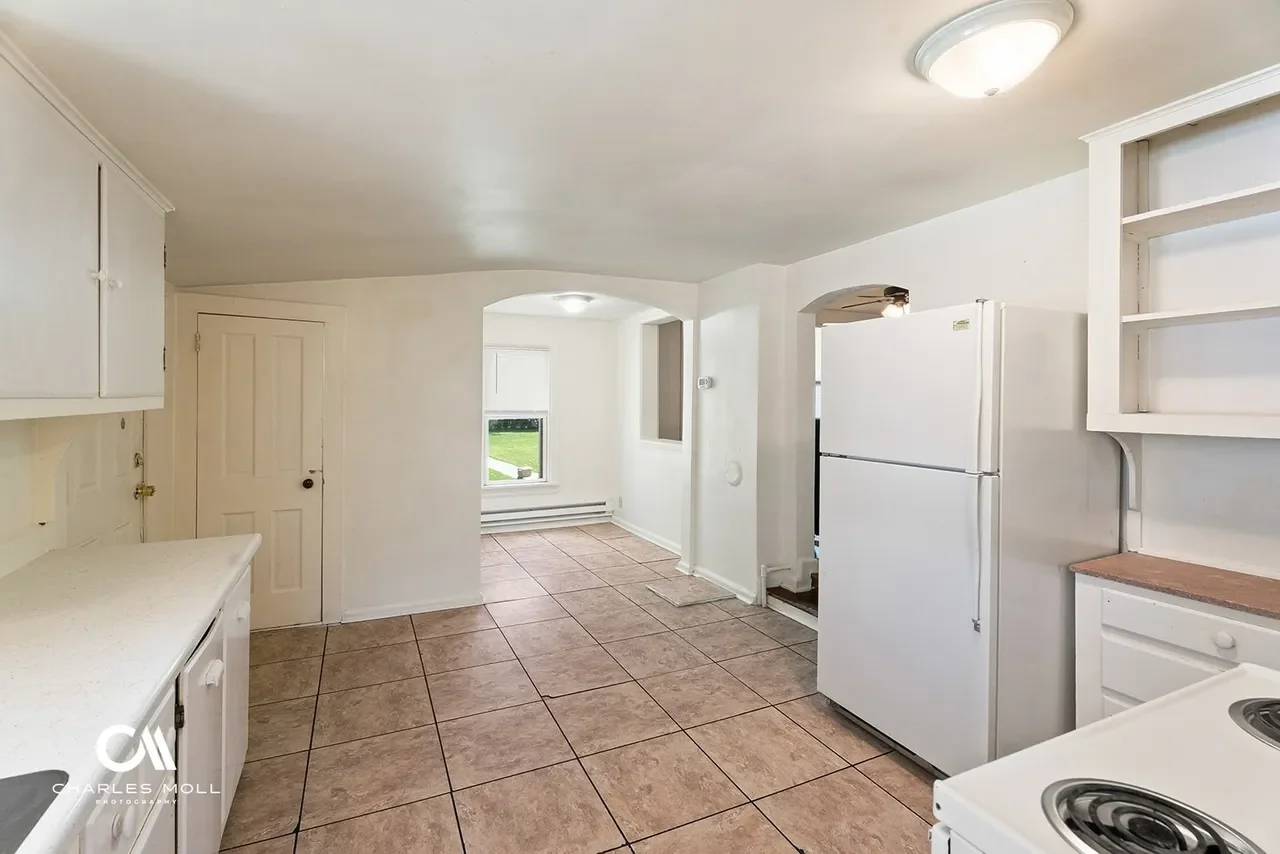 Empty kitchen with white cabinets, a white refrigerator, a stove, and a window overlooking a grassy yard.