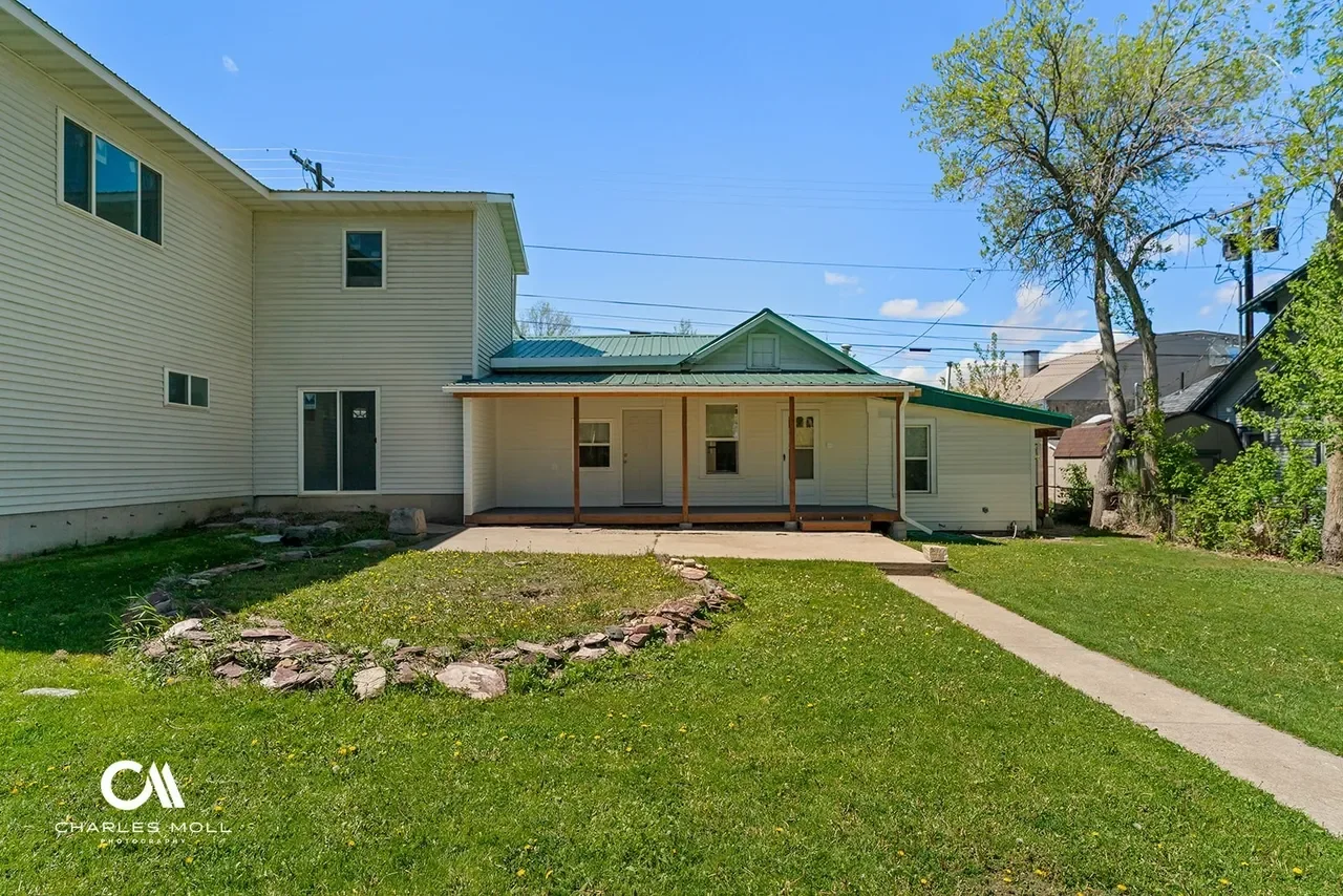 Backyard of a house with a grassy lawn, a stone-bordered garden bed, a concrete walkway, a small wooden porch with a green roof, and a large tree, under a bright blue sky.