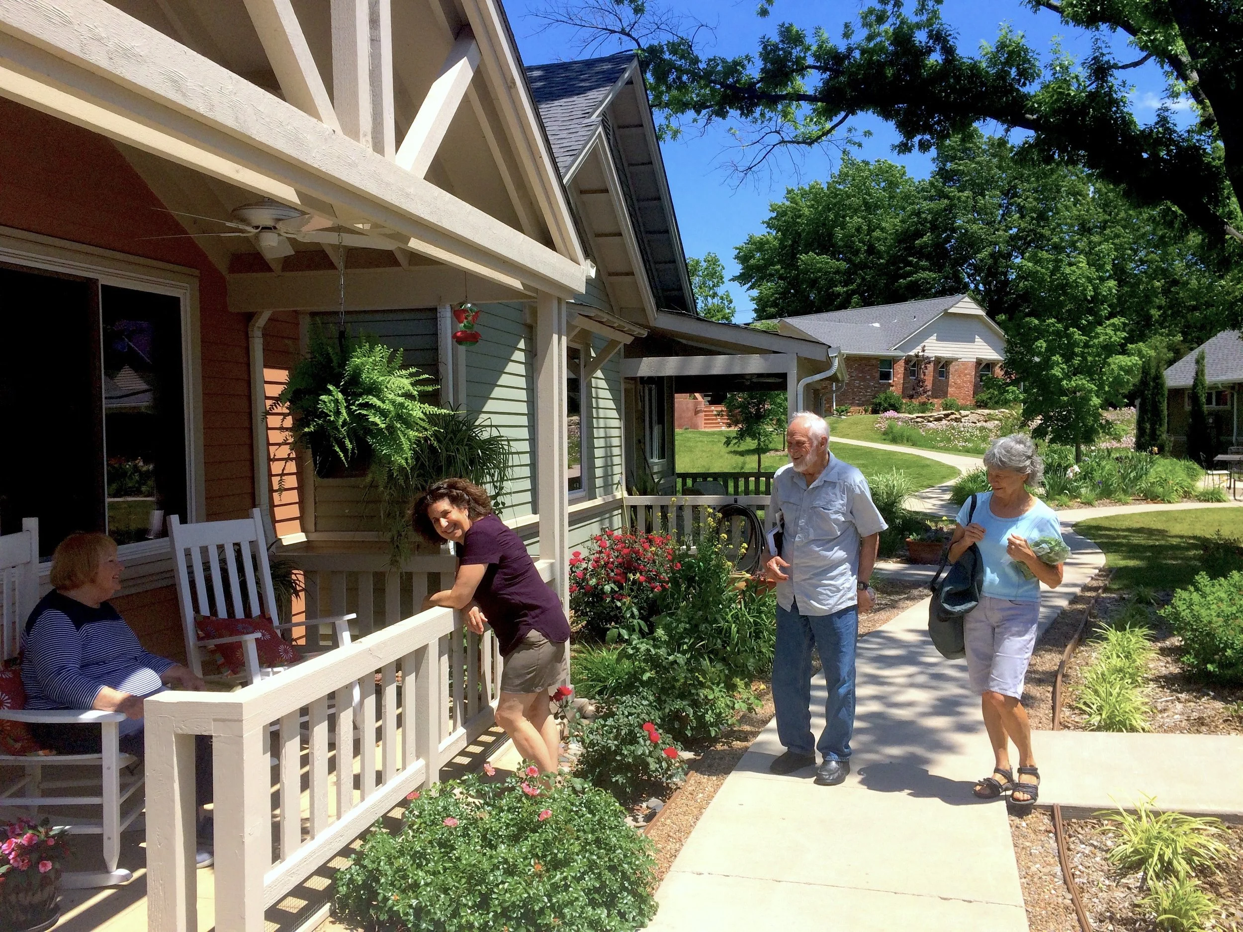 People gathered on a porch and sidewalk in a neighborhood on a sunny day. Four women and one man are visible, some sitting and some walking. The porch has white railings with potted plants, and the neighborhood has well-maintained houses with trees and landscaped yards.