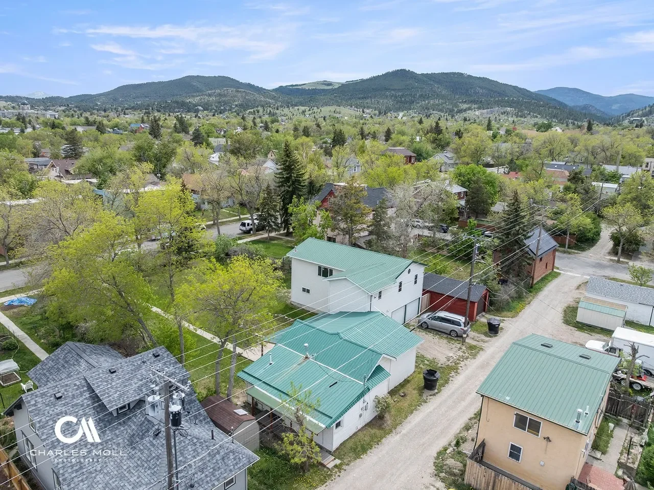 Aerial view of a residential neighborhood with houses, trees, and mountains in the background. Features include houses with green and gray roofs, dirt roads, and electrical poles.