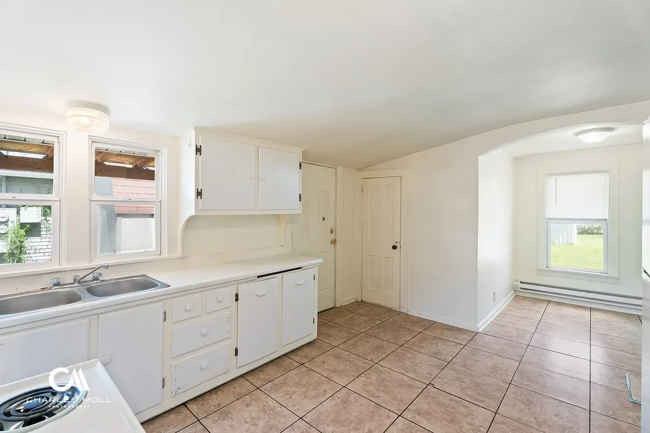Empty kitchen with white cabinets, a stainless steel double sink, four windows, tiled floor, and a small room with a window and baseboard heater.