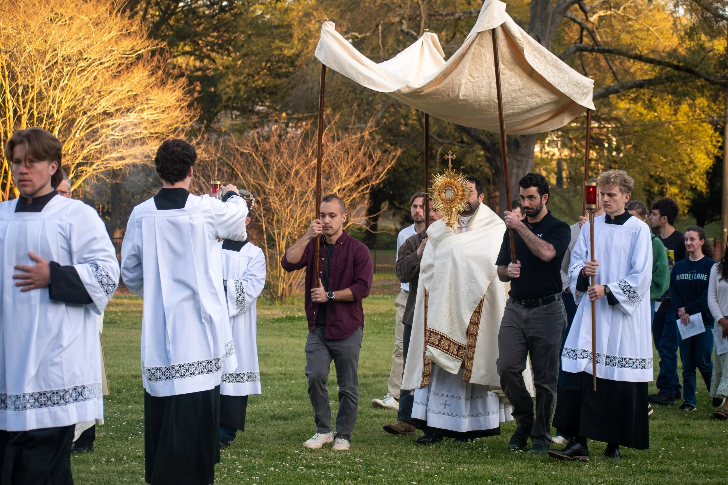 A religious outdoor ceremony with priests and altar servers holding a monstrance under a canopy, with onlookers and trees in the background during sunset.
