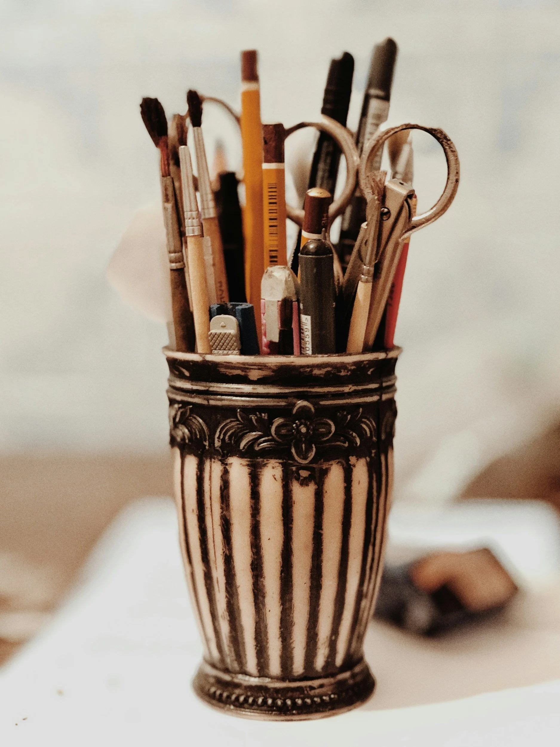 Close-up of a decorative metal vase filled with various pens and tools, placed on a white surface against a plain light background.
