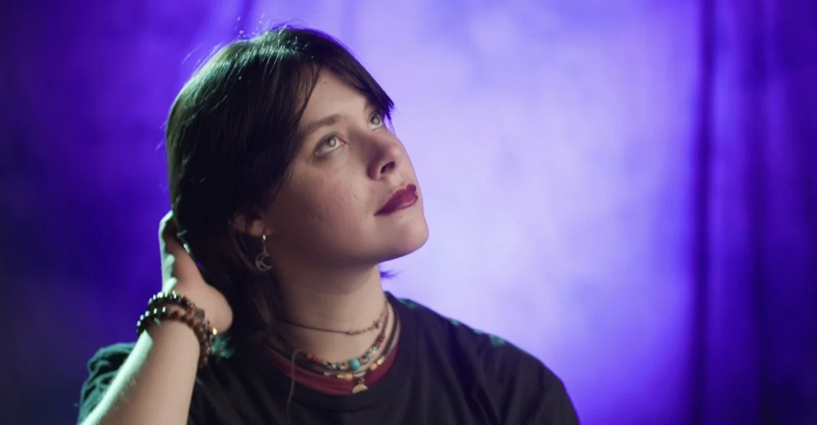 A young woman with short dark hair looking upwards, against a purple background.