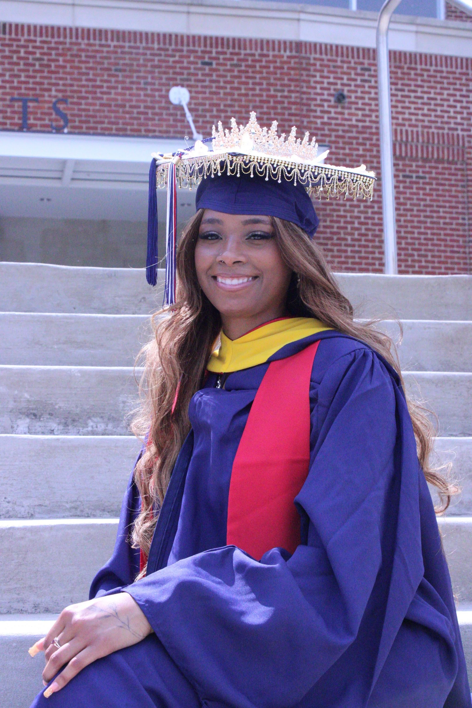 A young woman in graduation attire, wearing a blue cap and gown with a red and yellow hood, is sitting on outdoor steps. She has long wavy hair, a bright smile, and is wearing a tiara on her cap. The background features a brick wall and some structur