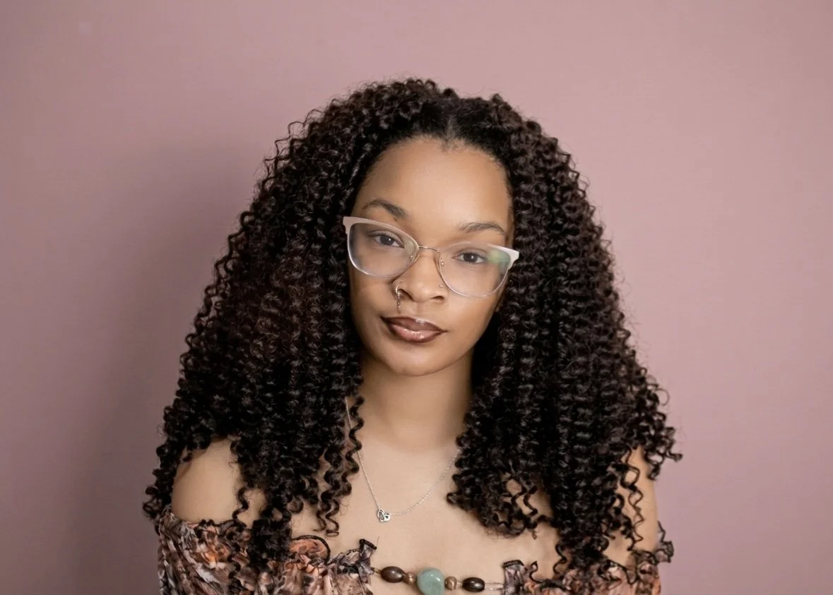 Portrait of a woman with curly hair, wearing glasses and jewelry, against a pink background.