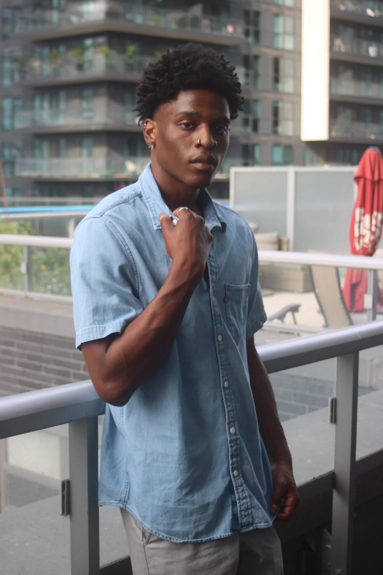 A young man with dark skin, short curly hair, wearing a light blue denim shirt and light-colored pants, standing outdoors on a balcony or terrace with modern buildings in the background. He has a serious expression, with his hand near his neck, and i