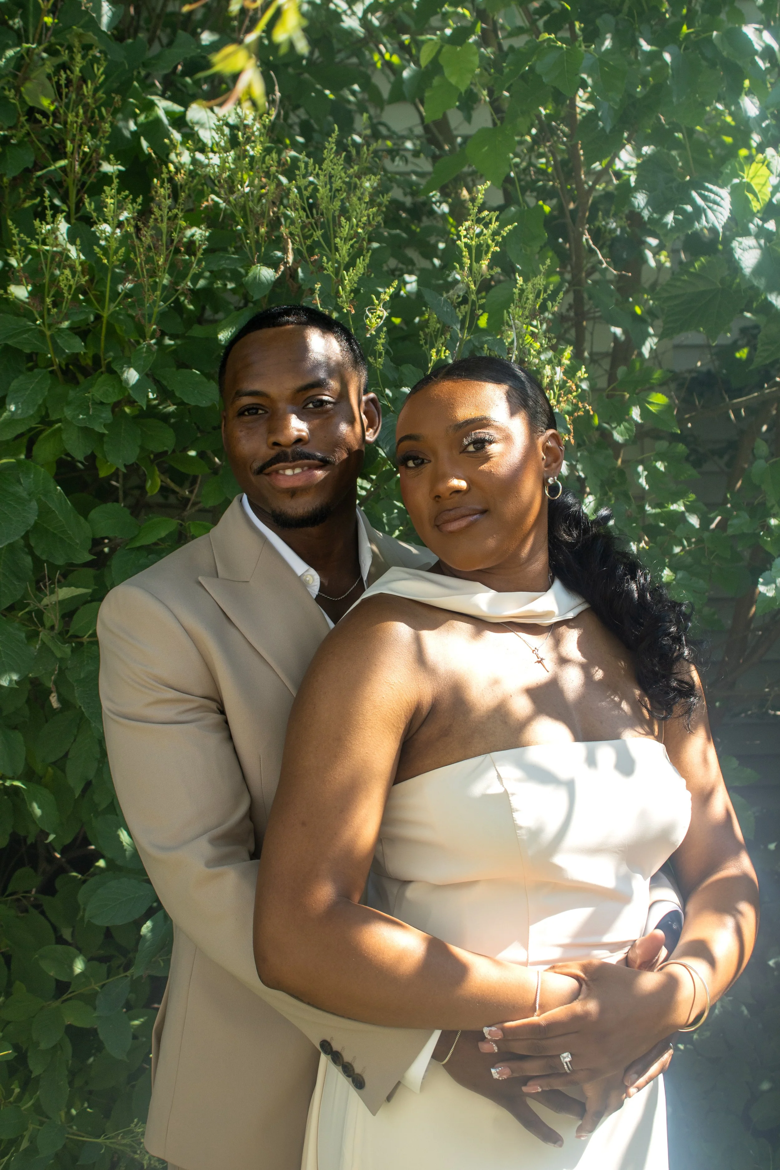 A couple dressed in formal white attire standing close together outdoors amidst green foliage, with the man's arm around the woman's waist, both smiling softly.