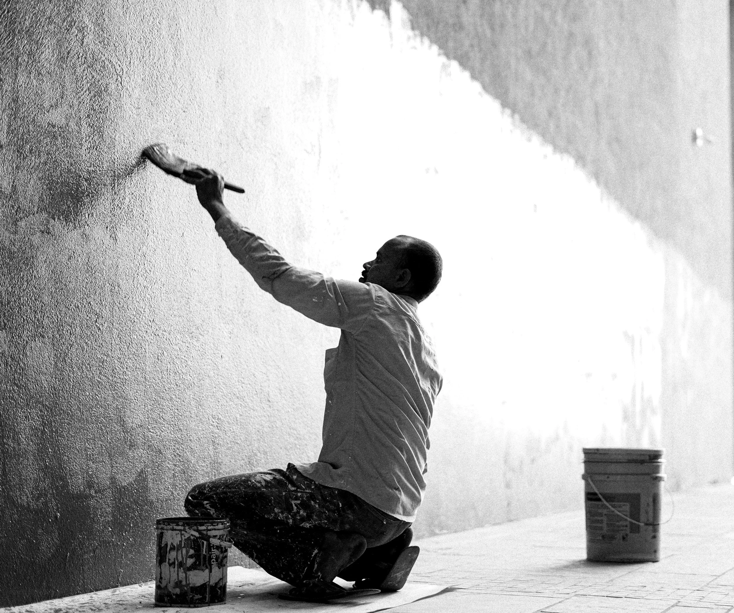 A person painting a wall with white paint while kneeling on the ground, with supplies and a large container nearby, outdoors on a sunny day.