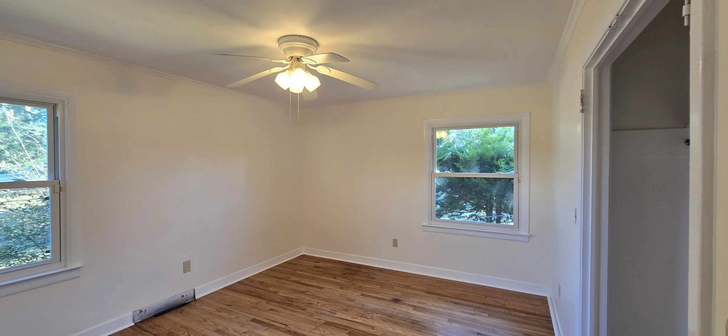 Empty room with hardwood floors, white walls, two windows showing greenery outside, a ceiling fan with lights, and an open doorway.