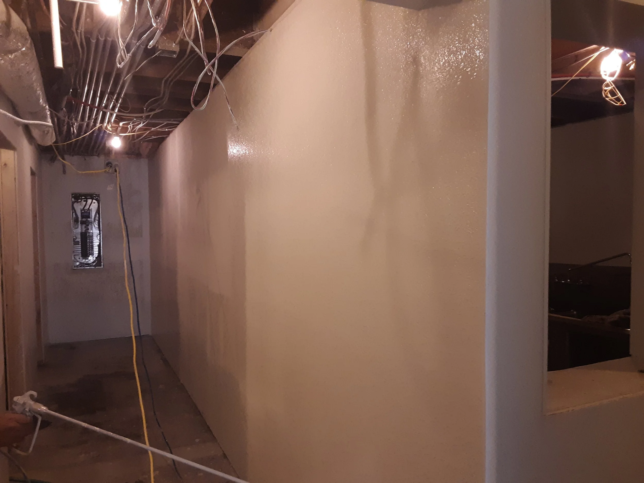 Unfinished basement with exposed ceiling beams, wiring, an electrical panel, and a freshly painted beige wall.