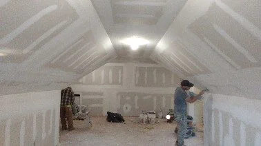 Two people working on drywall walls inside an unfinished attic or room with sloped ceilings.