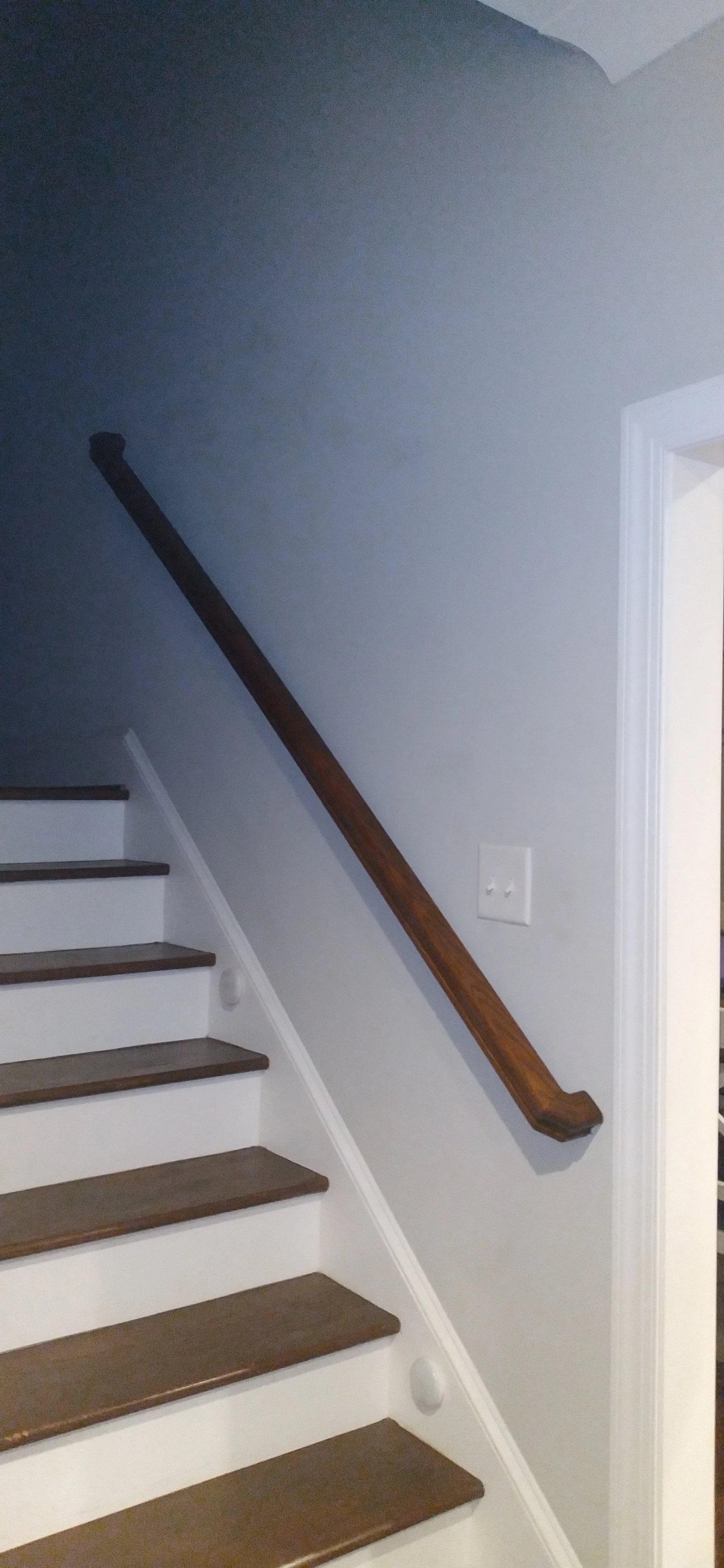 Photo of a staircase with wooden steps and a dark wooden handrail along a gray wall, next to a white door frame.