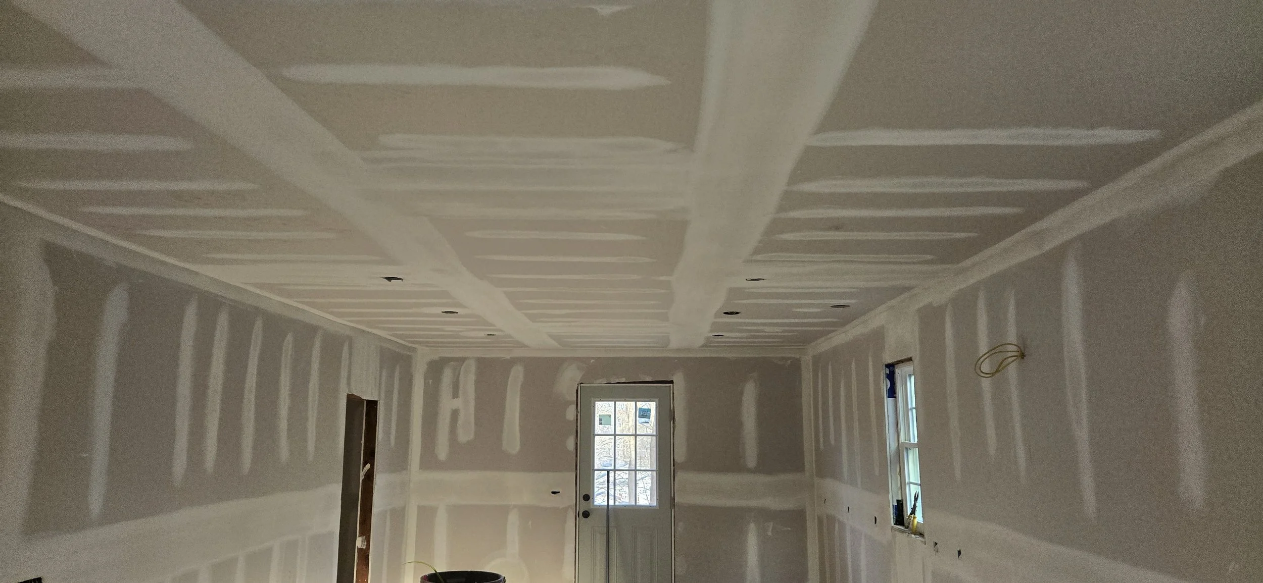 Interior room under construction with drywall installed on walls and ceiling, with visible joint compound markings, windows, and electrical wiring.