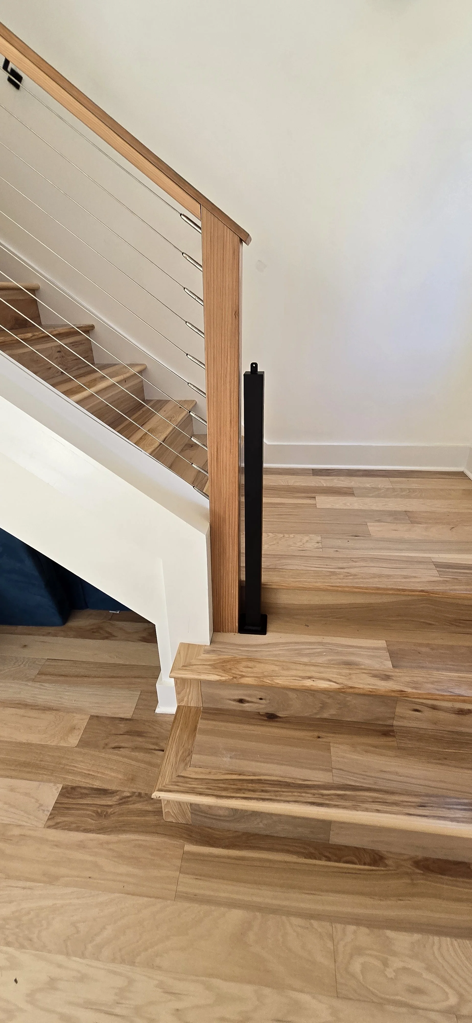 Interior view of a staircase with wooden steps, a wooden railing, and a white wall. There is a black vertical post at the corner of the staircase, and the flooring is wood.