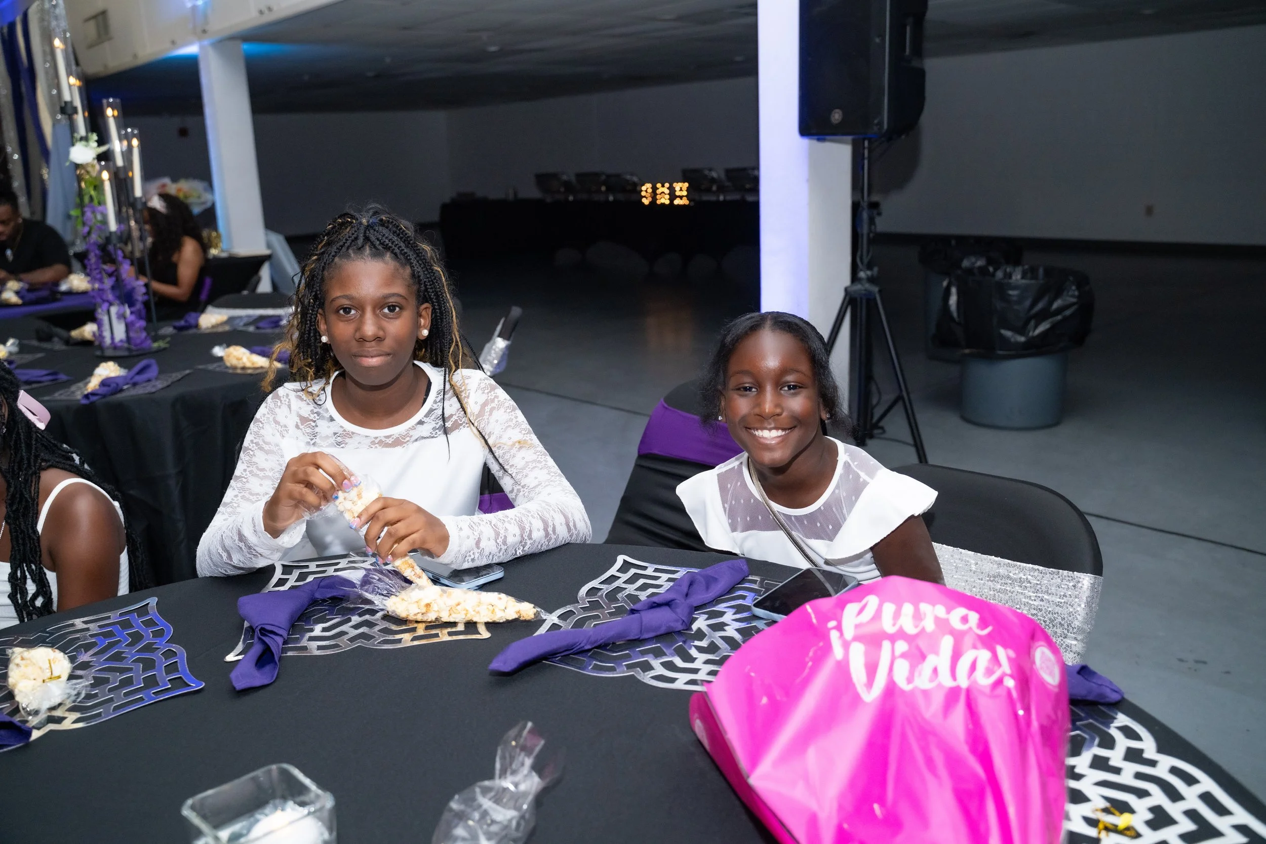 Two young girls sitting at a decorated table with purple and black themed tableware, eating slices of pizza during a celebration with a "Pure Birthday" balloon in the foreground.