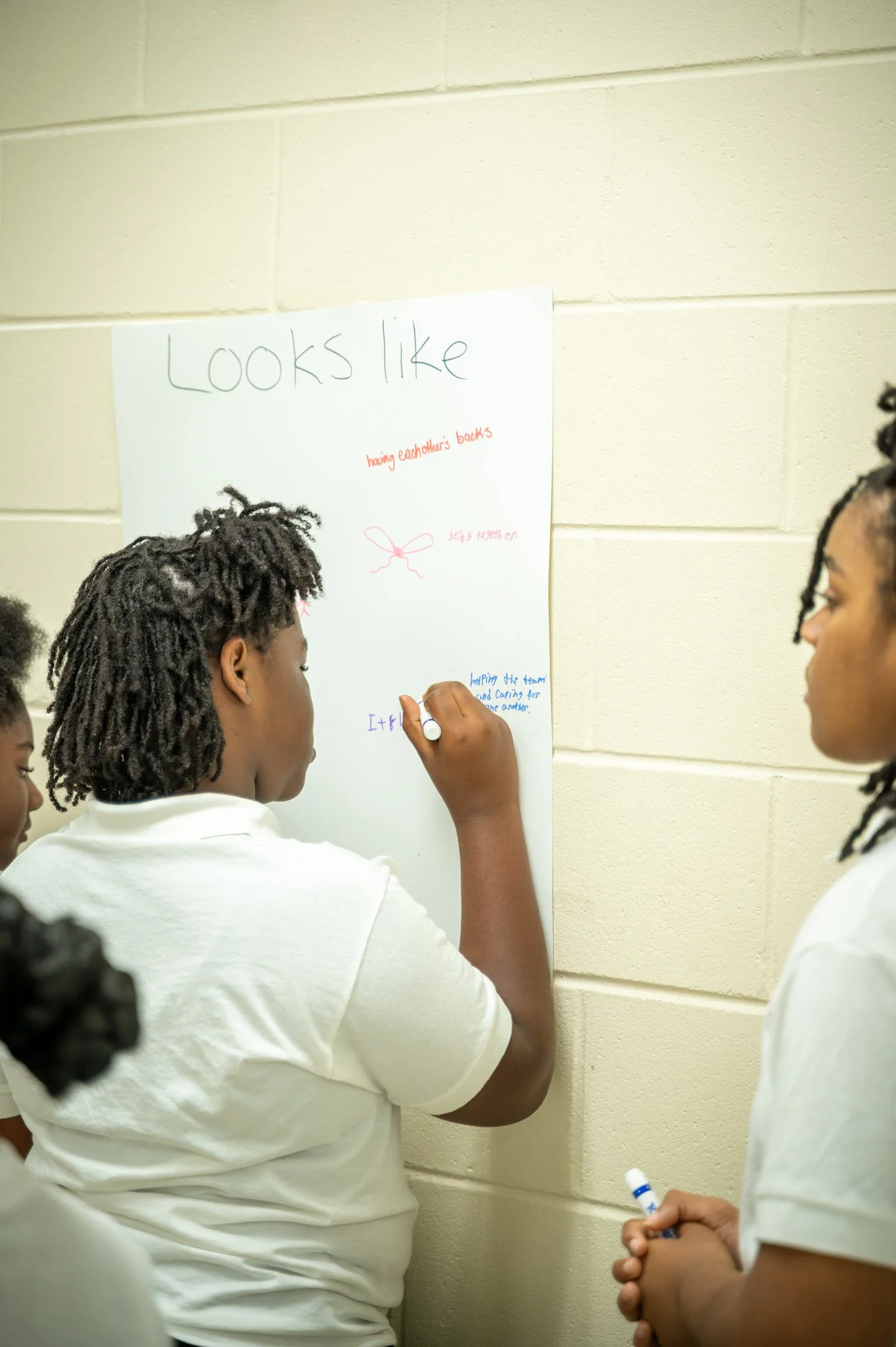 Students in a classroom writing on a poster that says 'Looks like' with handwritten notes and drawings.