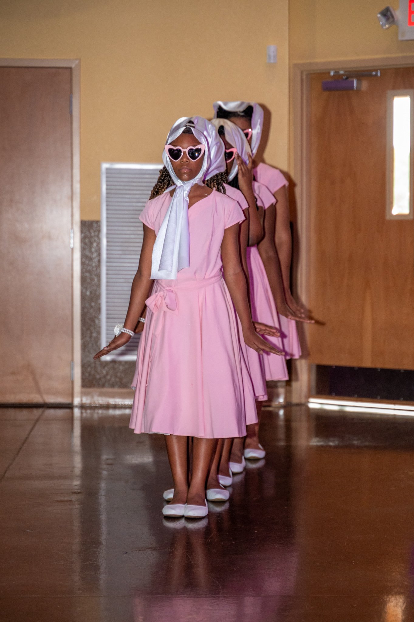 A line of young women dressed in matching pink dresses, wearing white shoes, and accessorized with white scarves and sunglasses, standing indoors in front of a wooden door and wall.