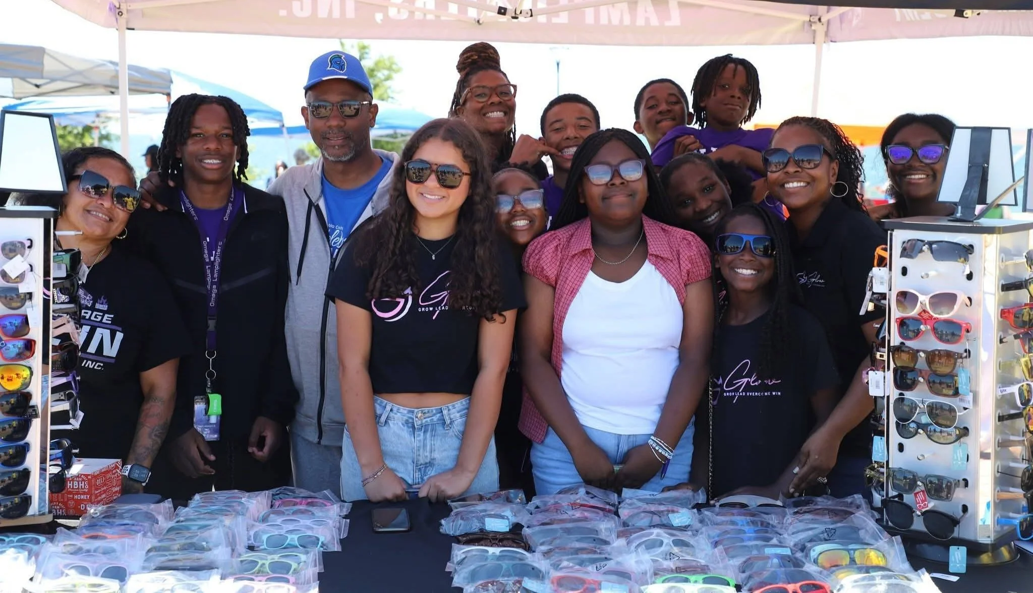 Group of diverse smiling people standing behind a sunglasses display booth at an outdoor event, under a canopy.