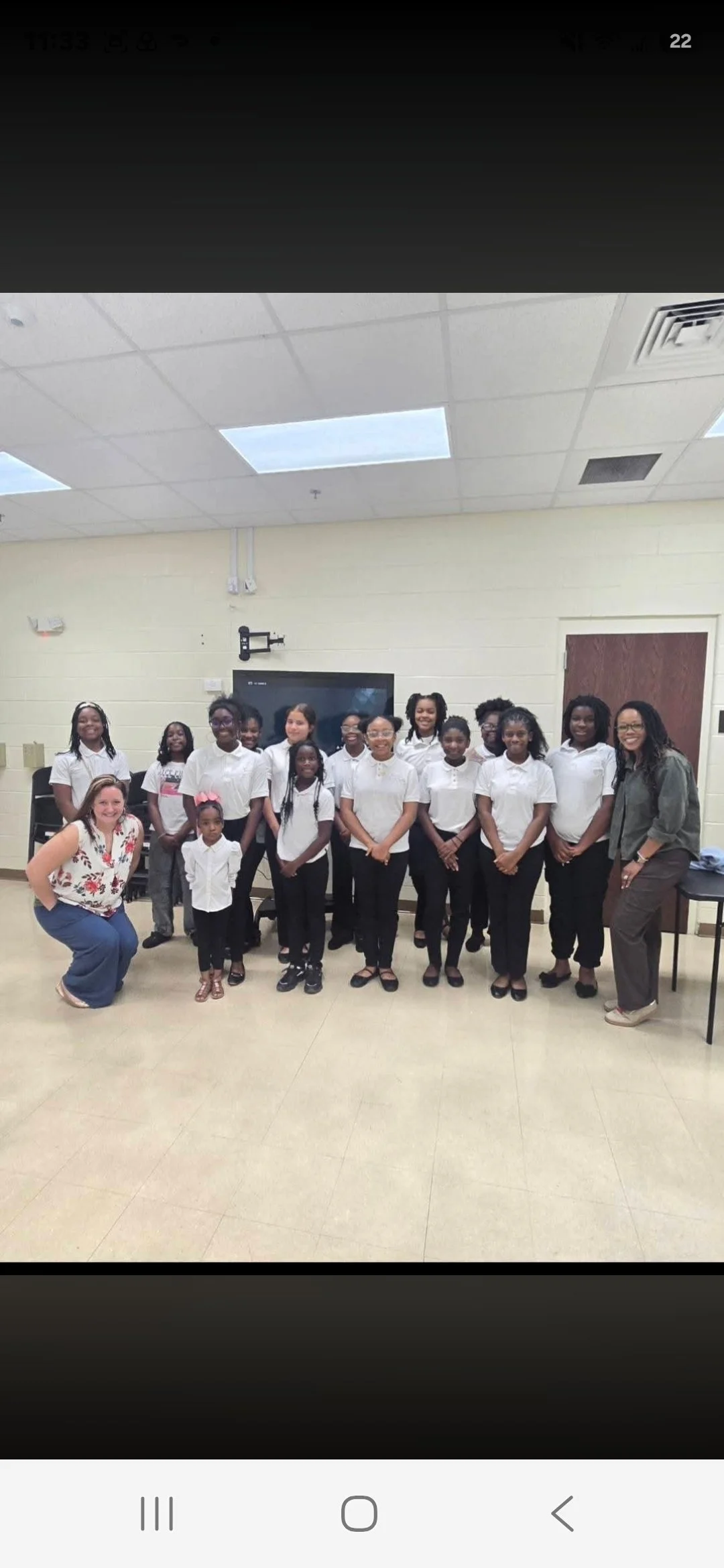 A group of young girls and two adult women smiling for a photo in a classroom or school setting, all dressed in coordinated white tops and dark pants.