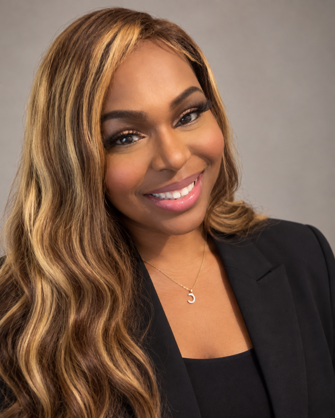 A woman with wavy, light brown hair and light brown skin, smiling, wearing a black blazer and a gold necklace with a letter 'C' pendant.