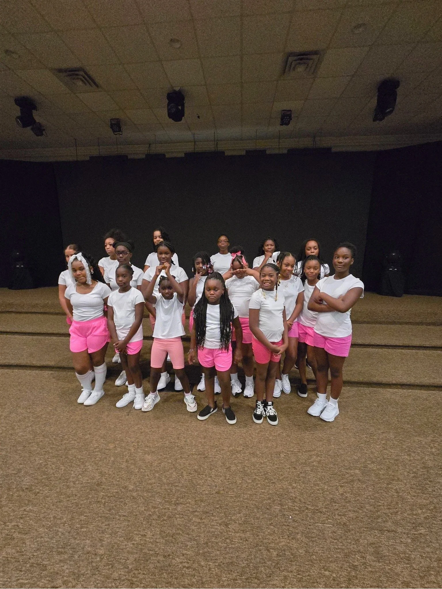 Group of young girls in white T-shirts and pink skirts standing on a stage in front of a black screen.