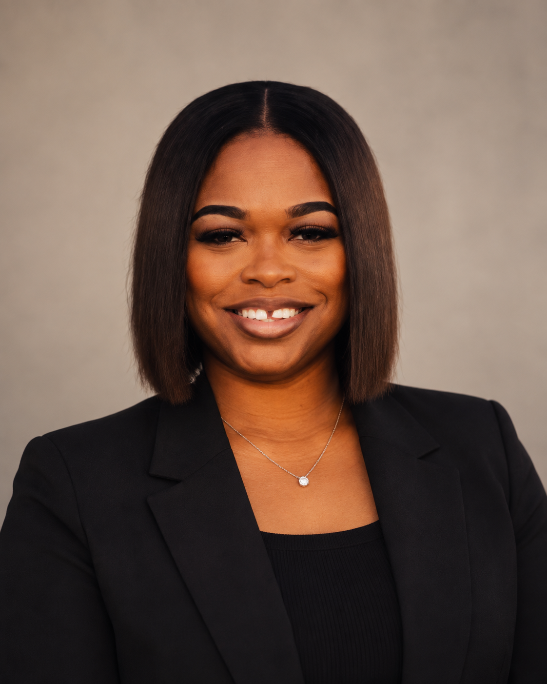 Portrait of a smiling woman with straight, shoulder-length hair, wearing a black blazer and a necklace, against a neutral background.