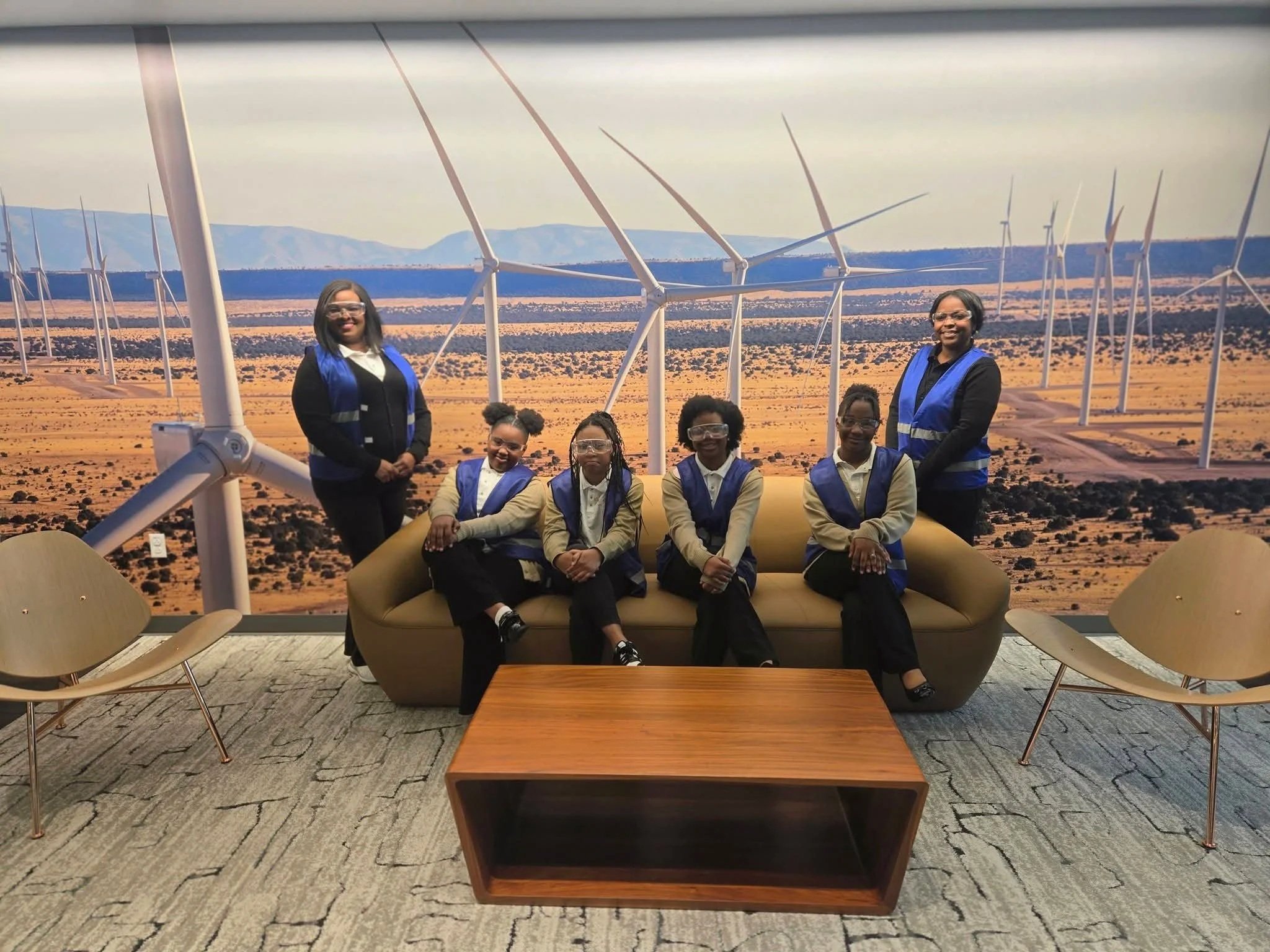 Group of six women wearing safety vests and glasses, sitting and standing around a mock-up wind turbine. Background is a large mural of a desert landscape with wind turbines and mountains.