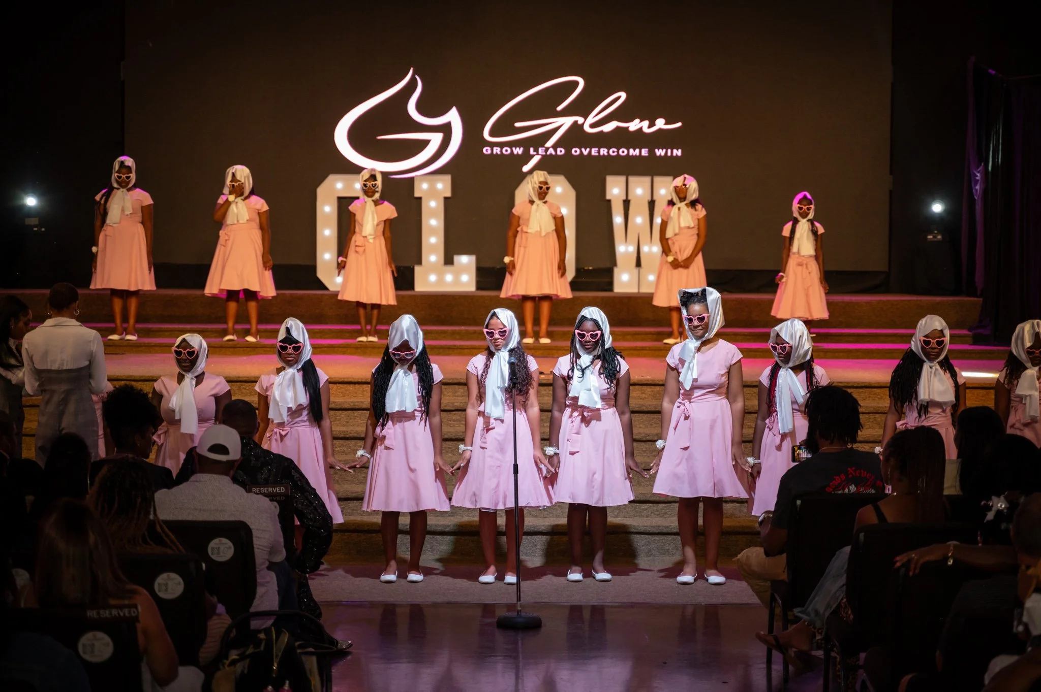 GLOW girls performing together in pink dresses on a stage with the GLOW logo.