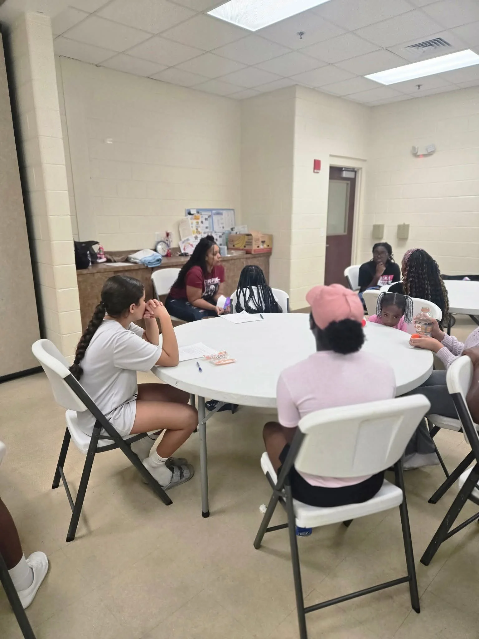 A group of teenagers sitting around a white round table in a classroom or community center. Some are talking, while others are listening. The room has beige walls and a dropped ceiling with fluorescent lights. A counter with various items is visible in the background.
