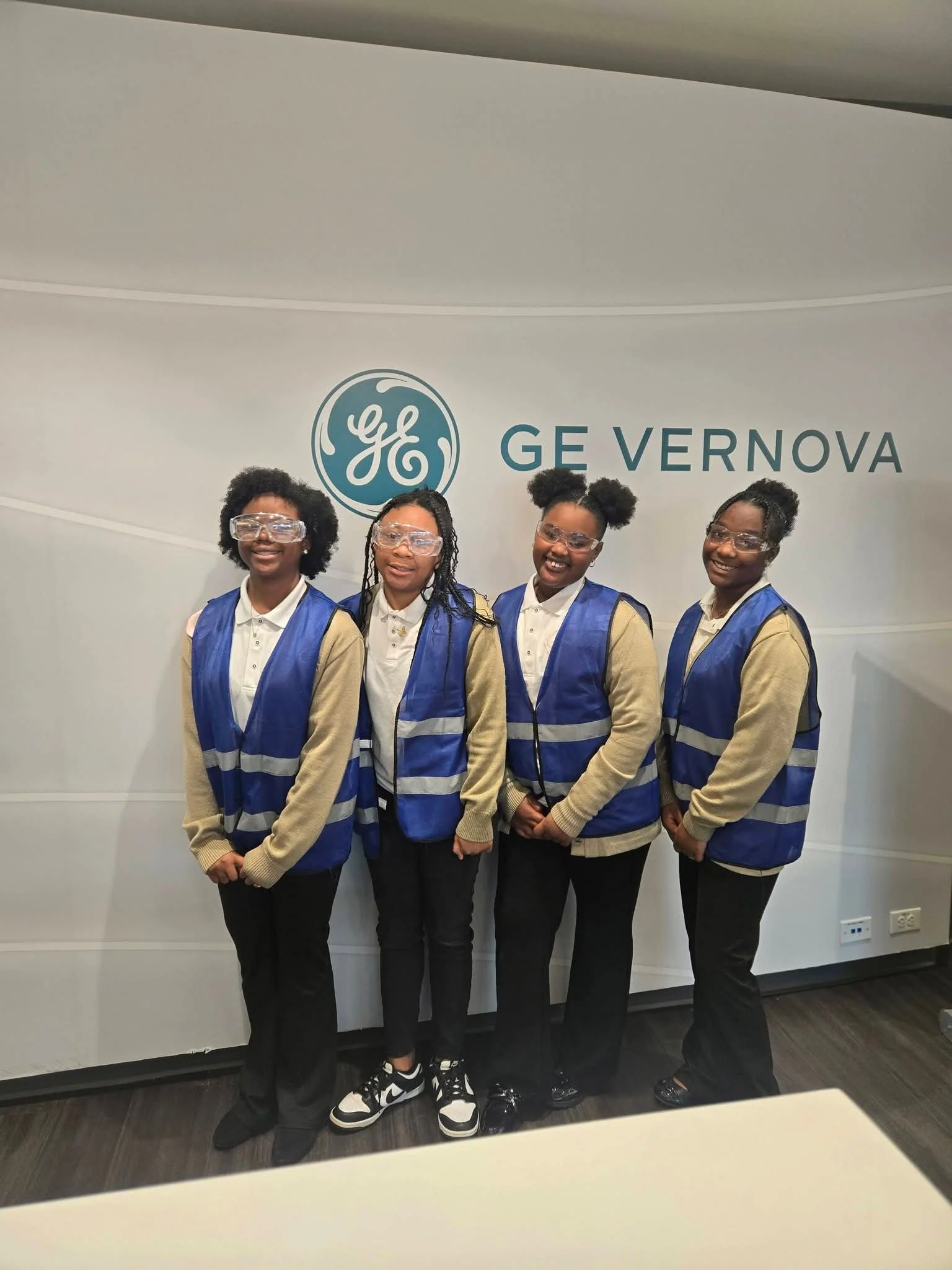 Four women standing together indoors, wearing safety glasses and blue vests, in front of a wall with the GE Vernova logo.