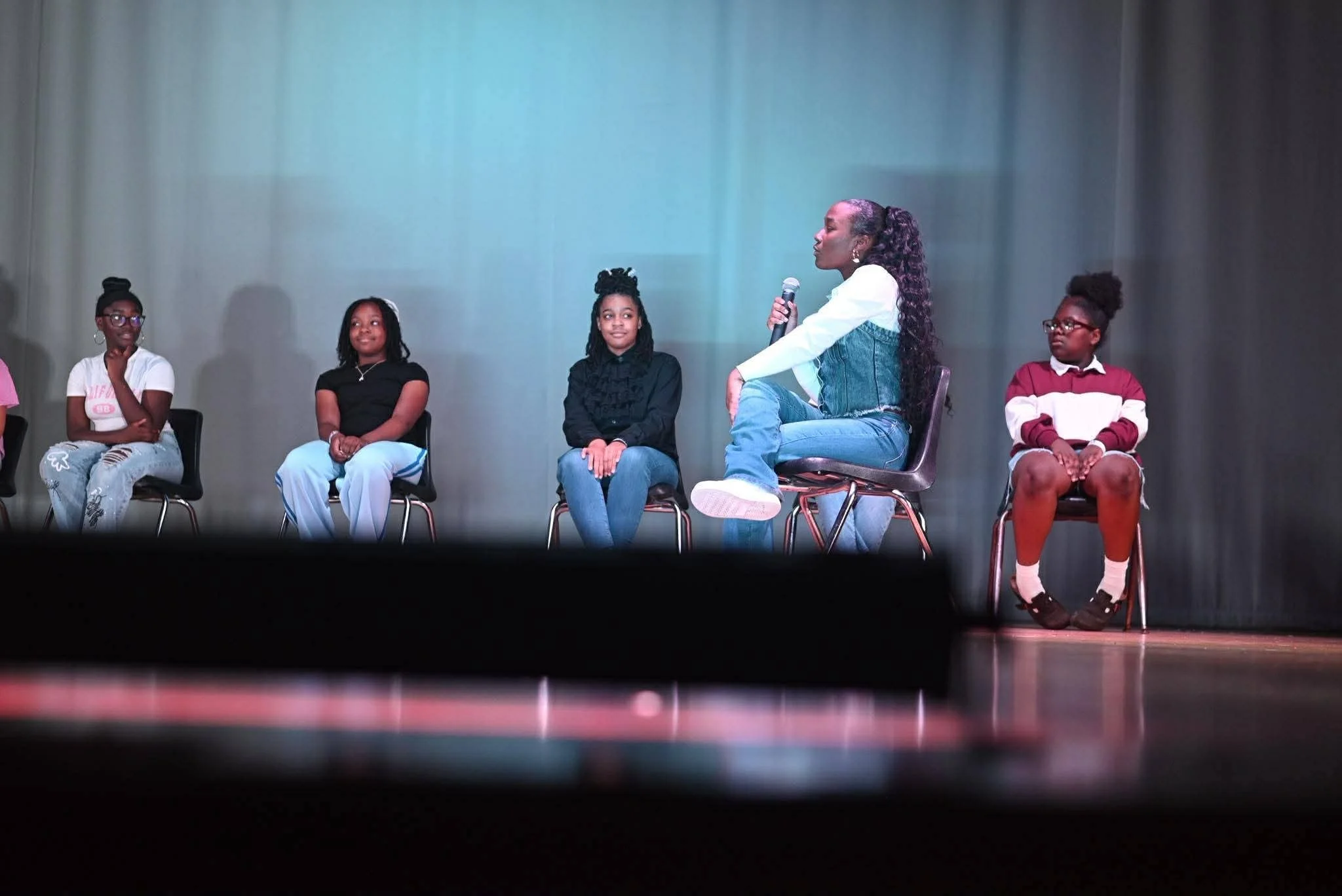 GLOW girls seated on stage while a speaker talks with a microphone.