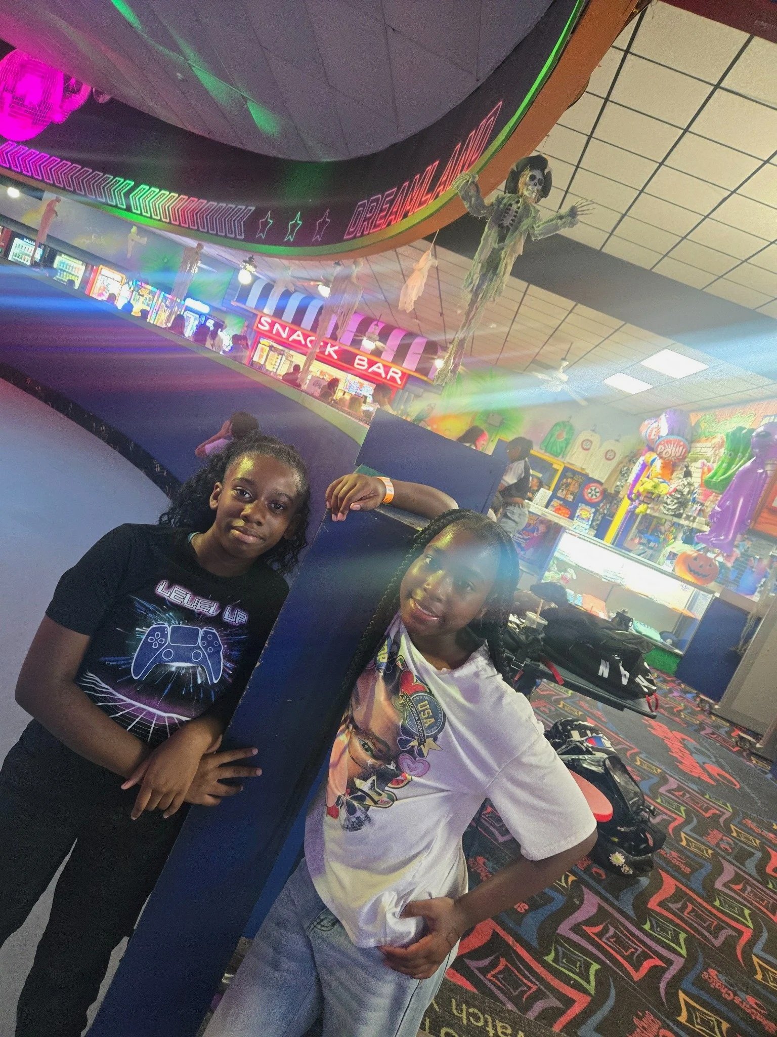 Two girls standing and posing at an indoor arcade or entertainment center, holding a large poster with space-themed artwork. There are neon lights, arcade machines, and Halloween decorations like hanging ghost figures in the background.