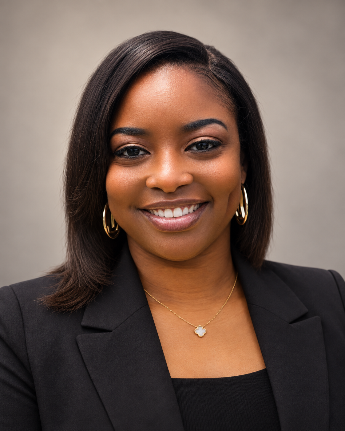 A smiling Black woman with shoulder-length dark hair, wearing a black blazer, gold hoop earrings, and a gold necklace with a four-leaf clover pendant, standing against a plain light gray background.