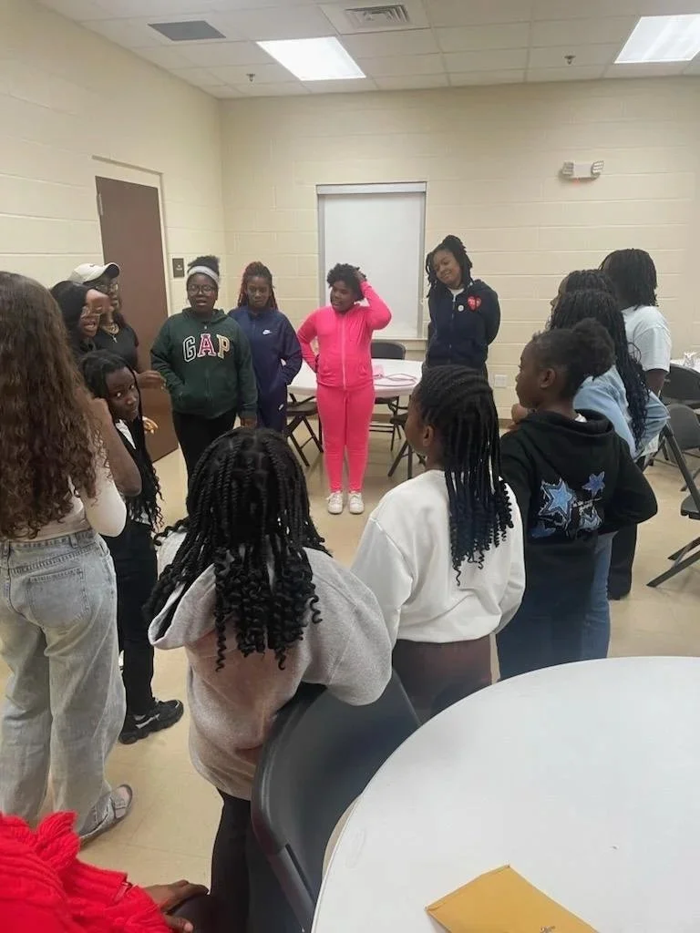 Group of young girls standing in a circle inside a room with beige walls and fluorescent lights, engaging in a discussion or activity.