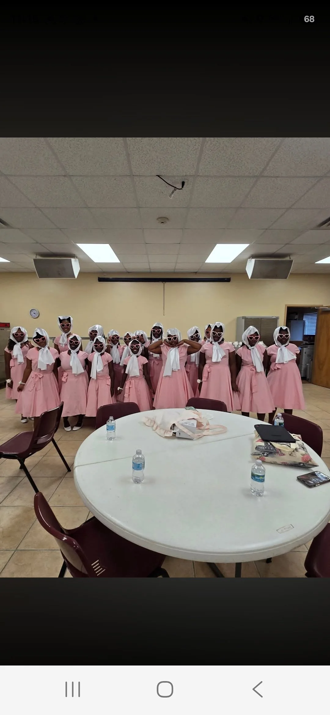 Group of women dressed in pink vintage-style dresses with white headscarves and white sunglasses, standing in a line in a room with beige walls, ceiling tiles, and tables with water bottles.